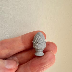 May include: A small, gray, acorn-shaped decorative object. The object has a textured surface and sits atop a pedestal base. The acorn is held in a person's hand against a neutral background.