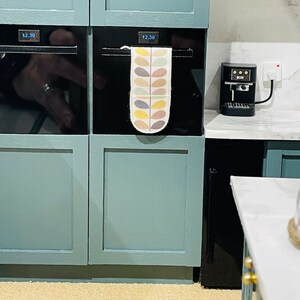 May include: A miniature kitchen with teal cabinets and two ovens, each displaying 12.30. A patterned tea towel hangs on one oven. A small black coffee machine is on the counter. The scene evokes a sense of a modern, compact kitchen.