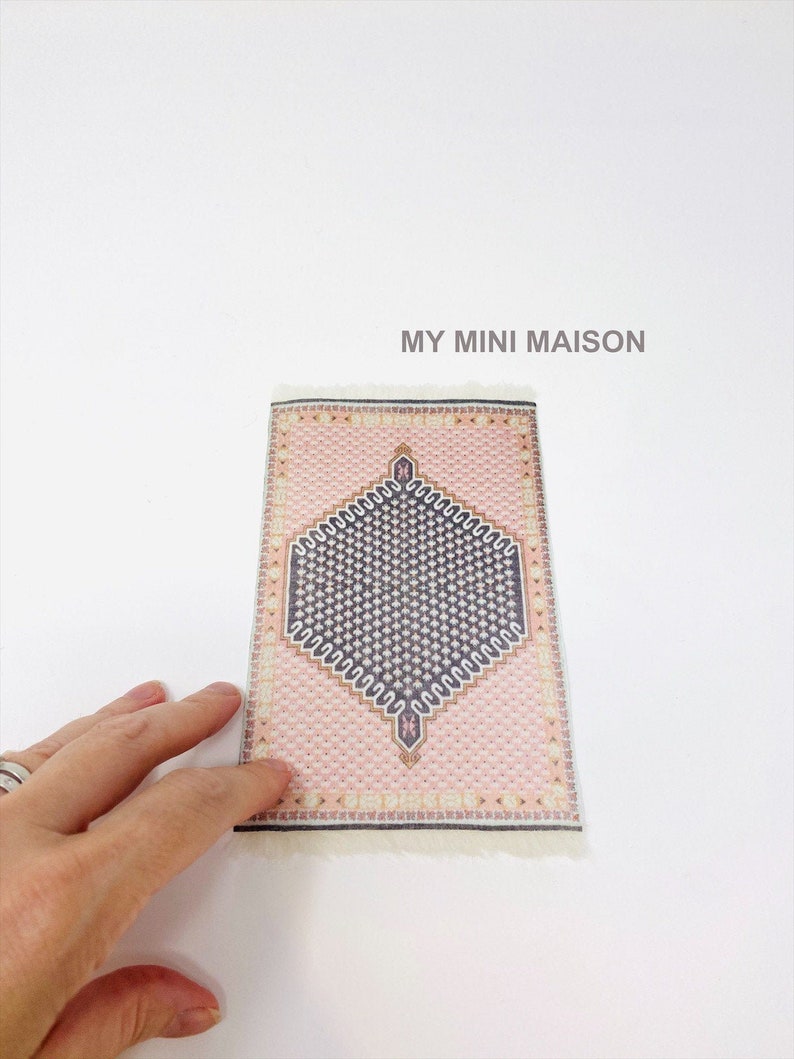 Pu&ograve; includere: Un tappeto rettangolare in miniatura con un motivo geometrico rosa e blu scuro. Il tappeto ha una frangia color crema lungo il bordo inferiore. Il testo "MY MINI MAISON" &egrave; stampato in grigio.