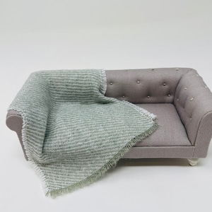 May include: Miniature grey sofa with button detailing and a light green and white patterned throw. The sofa has small white feet and is set against a white background. The blanket has a fringed edge.