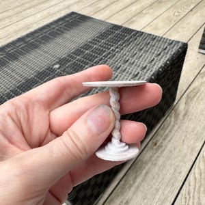 Puede incluir: Una mesa en miniatura de color blanquecino con un tallo retorcido y una parte superior e inferior redondas. La mesa se sostiene en una mano, mostrando su pequeño tamaño. La mesa mide aproximadamente 3,8 cm de alto y 2,5 cm de diámetro.