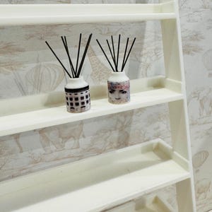 May include: Two white ceramic diffuser bottles with black reeds on a white shelf. One bottle has a black and white geometric pattern and the other has a colorful image of a woman's face.