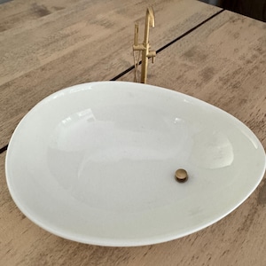 May include: A white, oval-shaped ceramic sink with a gold-colored faucet. The sink has a single drain hole with a gold-colored drain cover. The faucet is a minimalist design, with a curved spout and a single handle. The sink is set on a wooden surface.