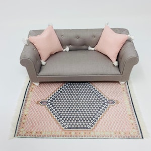 Pu&ograve; includere: Divano grigio in miniatura con due cuscini rosa e nappe bianche. Il divano &egrave; posizionato su un tappeto fantasia con un disegno geometrico rosa e blu navy. Il tappeto ha una frangia bianca.