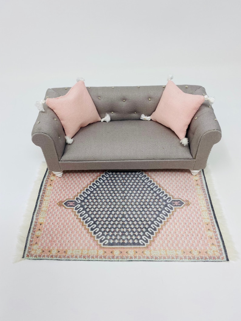 Pu&ograve; includere: Divano grigio in miniatura con due cuscini rosa e nappe bianche. Il divano &egrave; posizionato su un tappeto fantasia con un disegno geometrico rosa e blu navy. Il tappeto ha una frangia bianca.