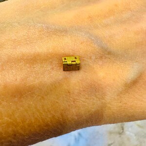 Op de afbeelding: Een klein, goudkleurig metalen scharnier. Het scharnier is gesloten en lijkt van messing te zijn.