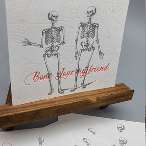 May include: A set of four white greeting cards with a skeleton design and the text "Bone Jour, my friend". The cards have a spiderweb pattern.