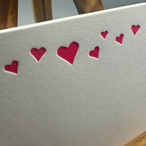 May include: A white card with a series of red heart cutouts arranged in a diagonal line. The hearts vary in size, with the largest heart at the bottom left and decreasing in size as they move up and to the right.