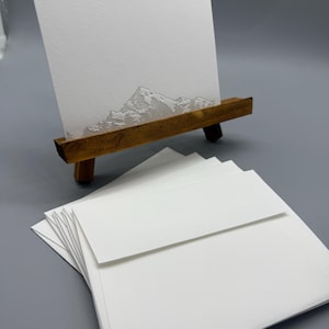 May include: A white card with an embossed mountain range design is displayed on a small wooden easel. A stack of matching white envelopes rests below. The background is a solid gray.