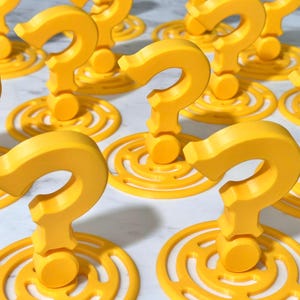 Könnte beinhalten: Eine Ansammlung leuchtend gelber Fragezeichen-Dekorationen. Jedes Fragezeichen ist 3D-gedruckt und steht auf einer kreisförmigen Basis mit konzentrischen Ringen. Die Objekte sind gleichmäßig verteilt und bilden ein Muster vor einem hellen Hintergrund. Das Gesamtthema deutet auf einen spielerischen oder lehrreichen Kontext hin.