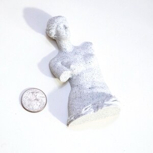 May include: A small, speckled, gray statue of a torso of a woman, with the head and upper body intact. The statue is next to a quarter dollar coin. The statue is approximately 4 inches tall.