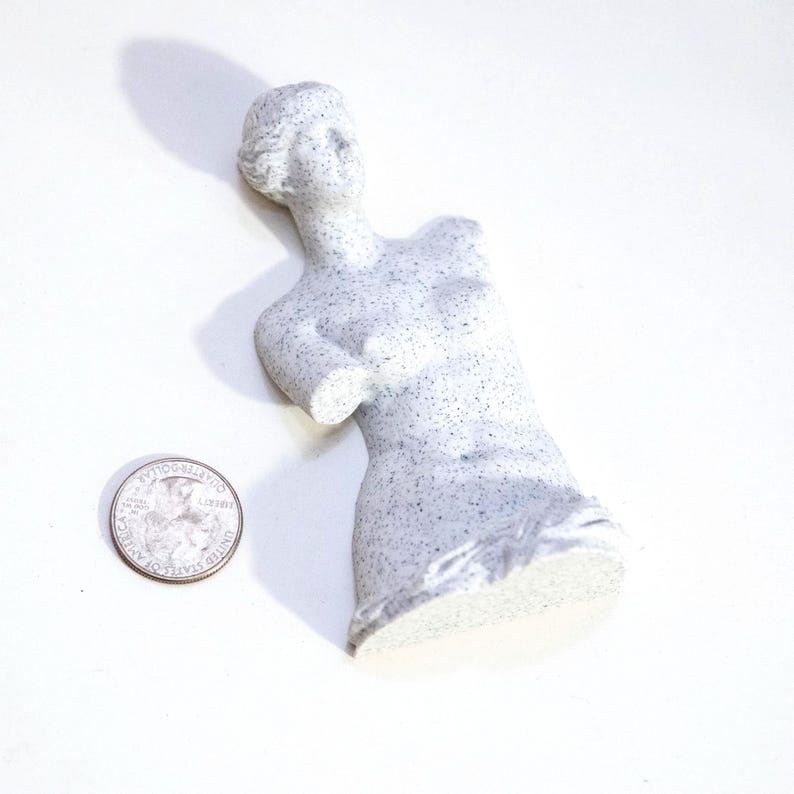 May include: A small, speckled, gray statue of a torso of a woman, with the head and upper body intact. The statue is next to a quarter dollar coin. The statue is approximately 4 inches tall.