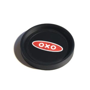 Puede incluir: Una tapa negra con un logotipo ovalado rojo y blanco que dice "OXO". La tapa redonda tiene un borde elevado y probablemente sea para un recipiente. La imagen está sobre un fondo blanco.