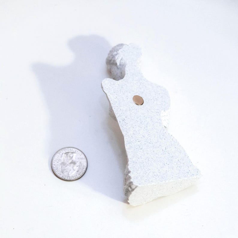 May include: A light gray, textured, abstract figurine with a small, round, copper-colored magnet attached. A U.S. quarter coin is next to the figurine for size comparison. The figurine has a rough, uneven edge.