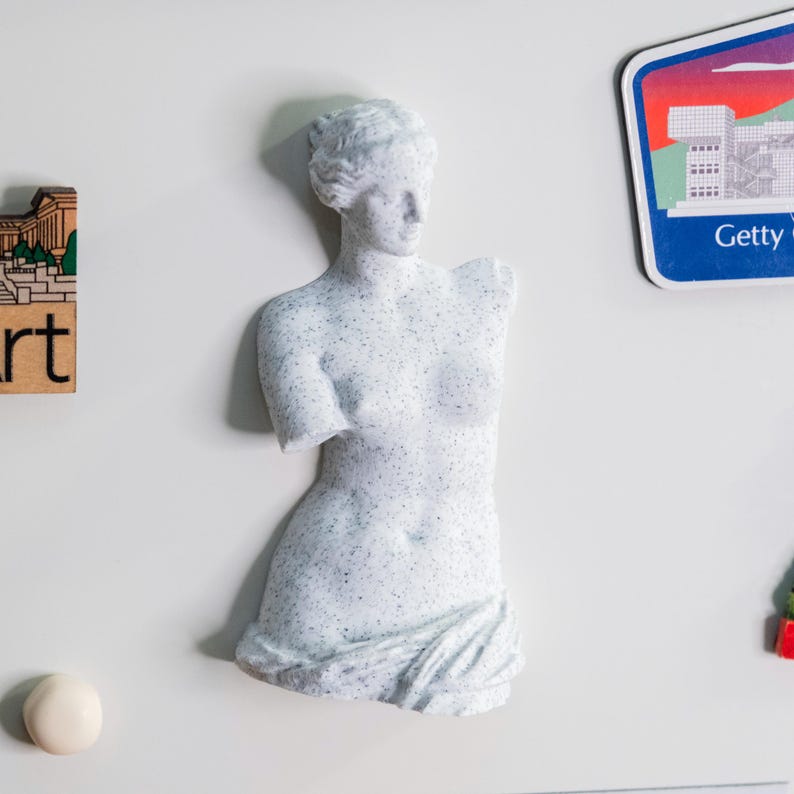May include: A white and speckled statue of a classical female figure, missing both arms. The sculpture is a bust, with detailed facial features and draped fabric around the lower body. The statue is a refrigerator magnet.
