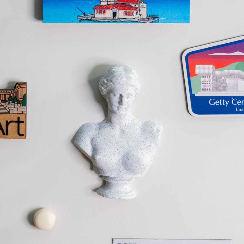 May include: A white, speckled bust of a classical female figure. The sculpture is mounted on a white surface, with other decorative items, including a magnet with the text "Getty Center Los" and a small painting of a building.