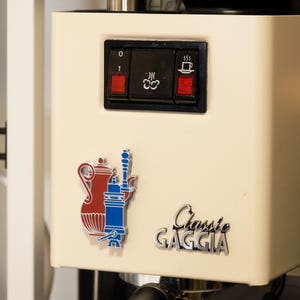 Peut inclure: Machine à expresso crème avec un panneau de commande noir avec des interrupteurs et des icônes rouges. Un graphique rouge et bleu et les mots "Classic Gaggia" sont sur le devant. La machine a une poignée noire et une base argentée.