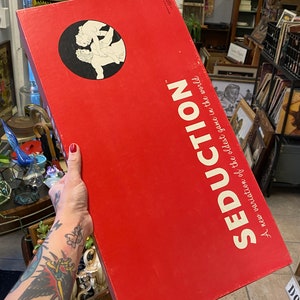 May include: A red box with the text "Seduction" in white. The text "A new variation of the oldest game in the world" is also printed on the box. The box has a black and white illustration of a couple on the front.