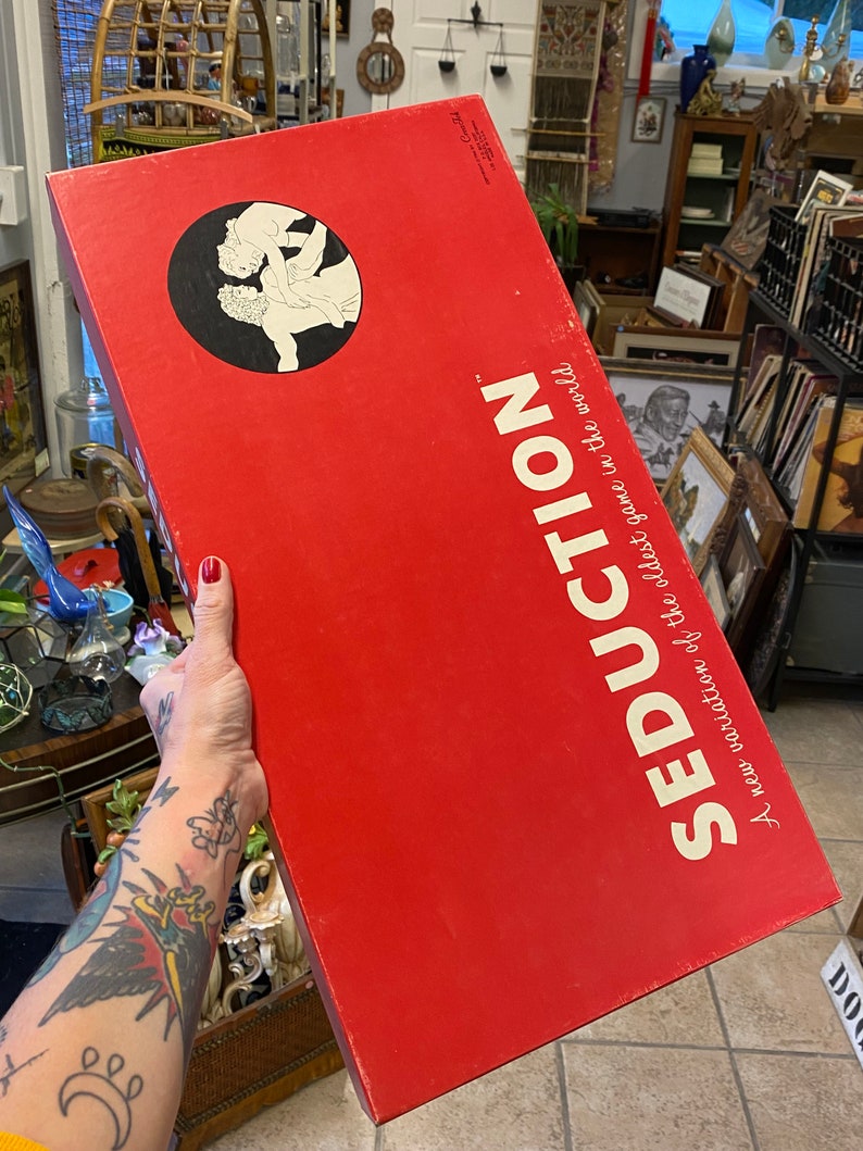 May include: A red box with the text "Seduction" in white. The text "A new variation of the oldest game in the world" is also printed on the box. The box has a black and white illustration of a couple on the front.