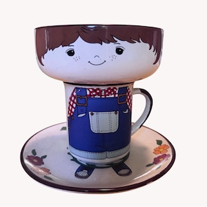 May include: A ceramic cup and saucer set featuring a cartoon character design. The cup depicts a figure in blue overalls and a red plaid shirt, while the bowl-shaped top shows a smiling face with brown hair. The saucer features small floral accents.