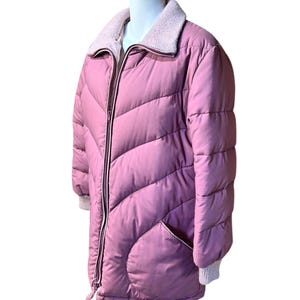 May include: A dusty rose puffer jacket with a light pink ribbed collar and cuffs. The jacket features a full zipper, two front pockets, and a drawstring at the hem. The quilted design adds texture to the outerwear.