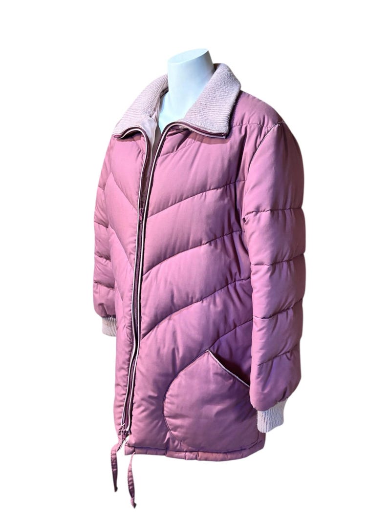 May include: A dusty rose puffer jacket with a light pink ribbed collar and cuffs. The jacket features a full zipper, two front pockets, and a drawstring at the hem. The quilted design adds texture to the outerwear.