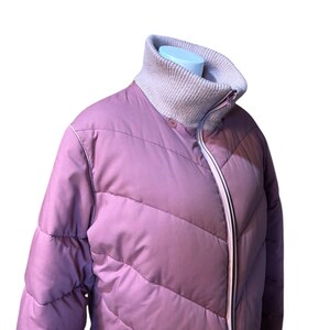 May include: A mauve puffer jacket with a high, ribbed collar and a full-length zipper. The jacket has a quilted design with diagonal stitching and a light pink trim. The collar is a light gray color.
