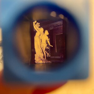 May include: A close-up view of a vintage photograph through a magnifying glass. The photo shows a white marble sculpture of two figures, a woman holding a branch and a man standing beside her.