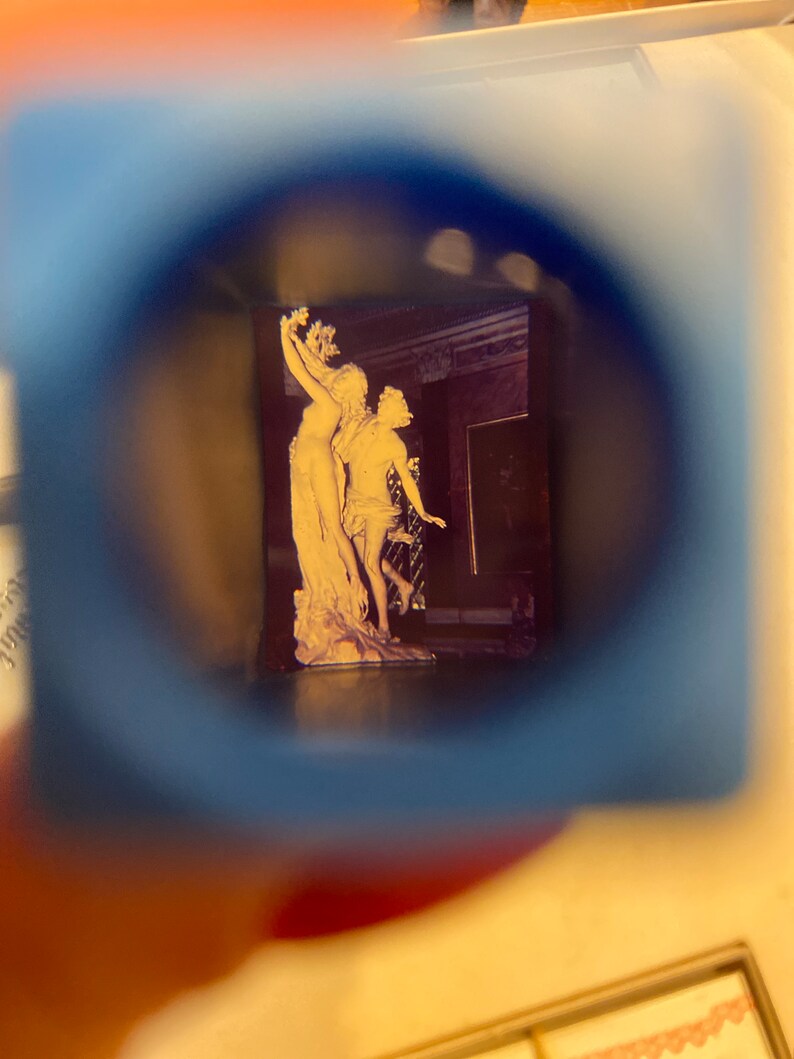 May include: A close-up view of a vintage photograph through a magnifying glass. The photo shows a white marble sculpture of two figures, a woman holding a branch and a man standing beside her.