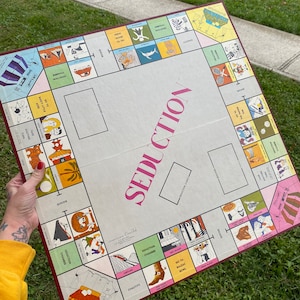 May include: A vintage board game called "Seduction" with a pink and white design. The board features illustrations of people, animals, and objects. The game is set on a white background with a red border.