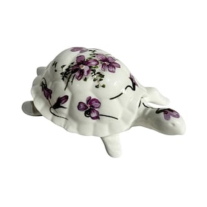May include: A white ceramic turtle figurine with a shell decorated with purple floral designs. The turtle has four legs and a head, all in white. The floral pattern includes purple flowers and green leaves.