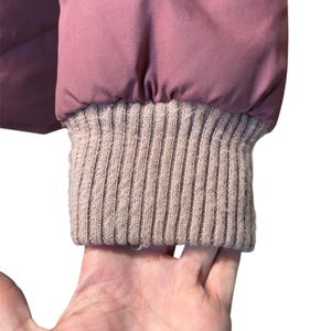 May include: Close-up of a mauve puffer jacket sleeve with a ribbed, light brown knit cuff. The jacket's fabric appears smooth and slightly shiny. The cuff is a contrasting texture to the jacket.