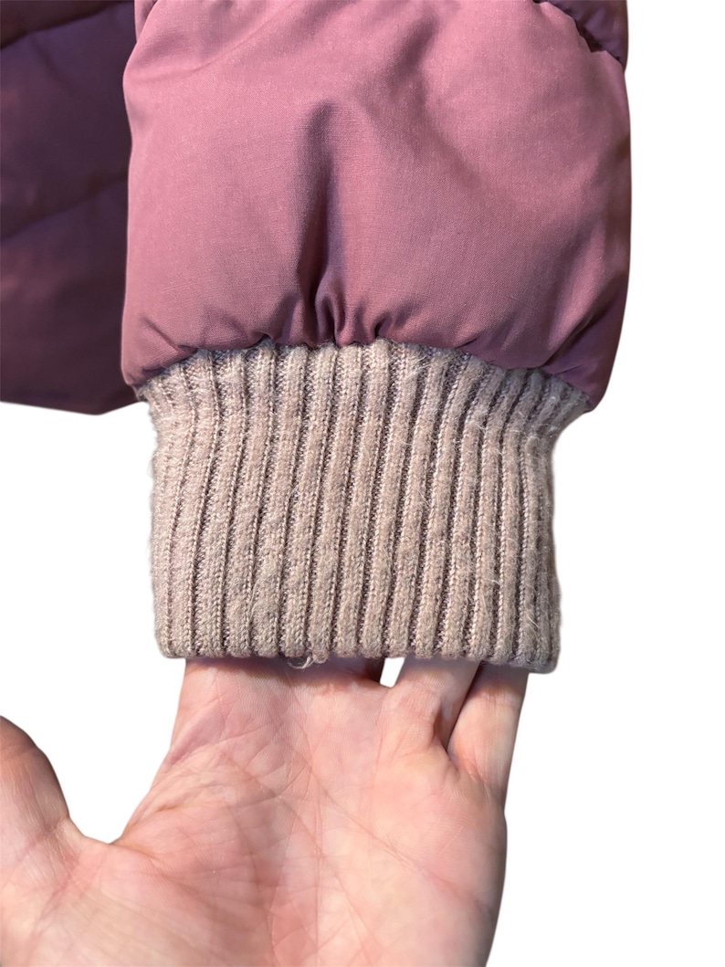 May include: Close-up of a mauve puffer jacket sleeve with a ribbed, light brown knit cuff. The jacket's fabric appears smooth and slightly shiny. The cuff is a contrasting texture to the jacket.