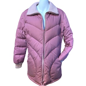 May include: A mauve-colored, quilted puffer jacket with a ribbed collar and cuffs. The jacket features a full-length zipper, two front pockets, and a drawstring at the hem. The jacket is displayed on a mannequin.