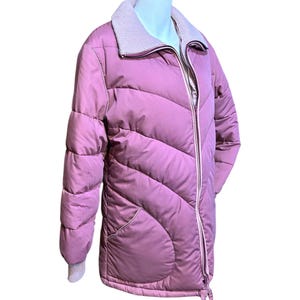 May include: A mauve-colored, quilted puffer jacket with a light gray knit collar and cuffs. The jacket features a full-length zipper, two front pockets, and a drawstring at the hem. The jacket is displayed on a mannequin.