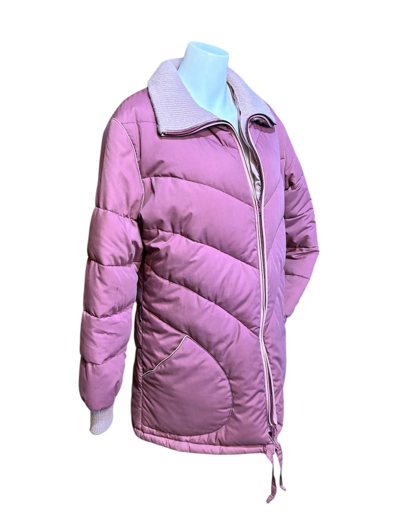 May include: A mauve-colored, quilted puffer jacket with a light gray knit collar and cuffs. The jacket features a full-length zipper, two front pockets, and a drawstring at the hem. The jacket is displayed on a mannequin.