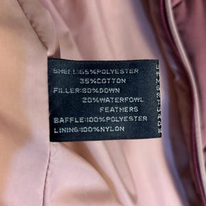 May include: Close-up of a black clothing tag with white text. The tag lists the shell as 65% polyester and 35% cotton. The filler is 80% down and 20% waterfowl feathers. The baffle is 100% polyester, and the lining is 100% nylon.
