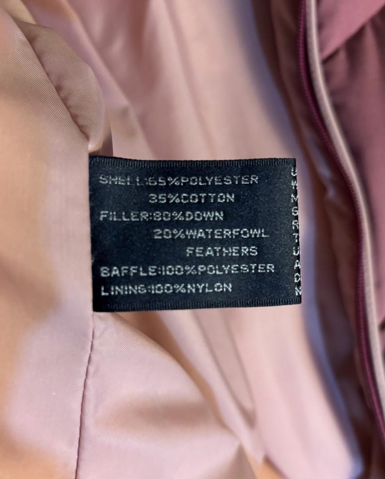 May include: Close-up of a black clothing tag with white text. The tag lists the shell as 65% polyester and 35% cotton. The filler is 80% down and 20% waterfowl feathers. The baffle is 100% polyester, and the lining is 100% nylon.