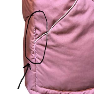 May include: Close-up of a dusty rose-colored quilted garment. The garment has a pocket with white trim. The stitching is visible, and the fabric appears soft and padded. The overall design is simple and functional.