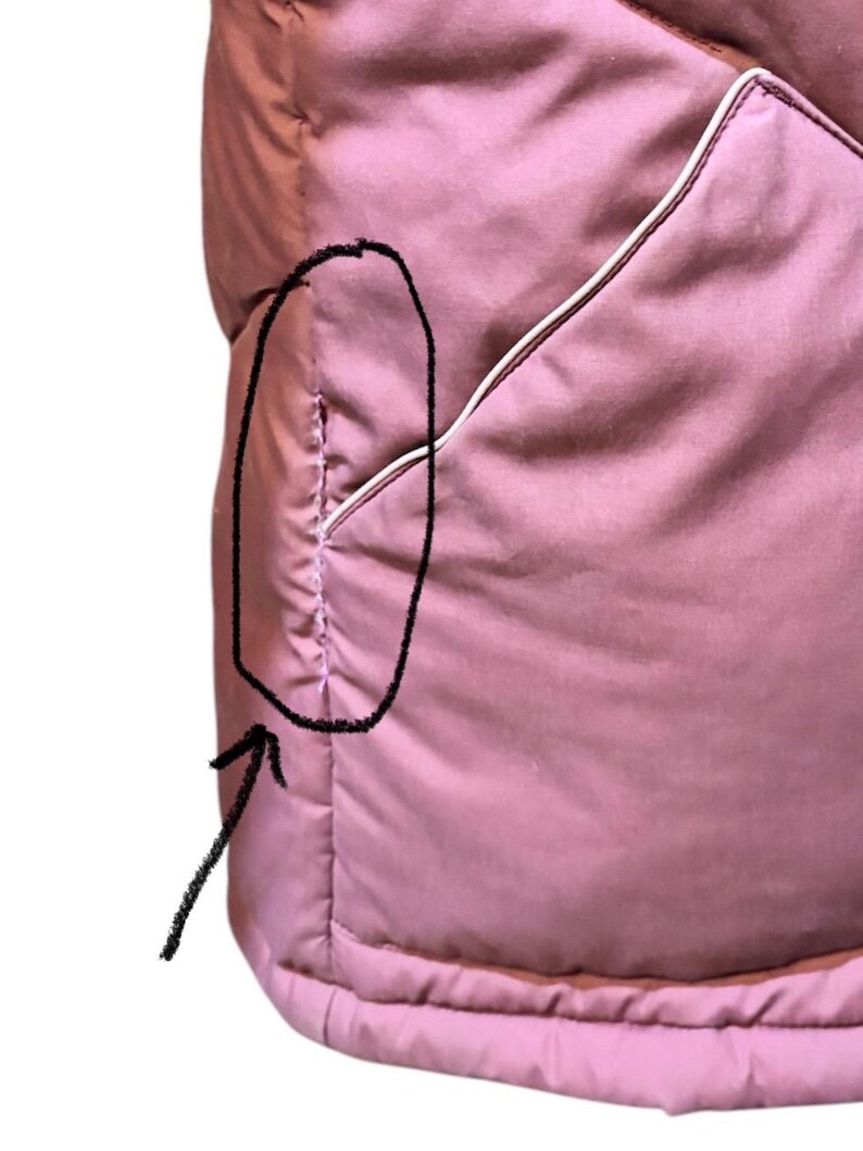 May include: Close-up of a dusty rose-colored quilted garment. The garment has a pocket with white trim. The stitching is visible, and the fabric appears soft and padded. The overall design is simple and functional.