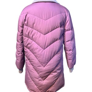 May include: A long, quilted, mauve-colored coat with a chevron design. The coat has a light gray knit collar and cuffs. The coat is displayed on a mannequin.