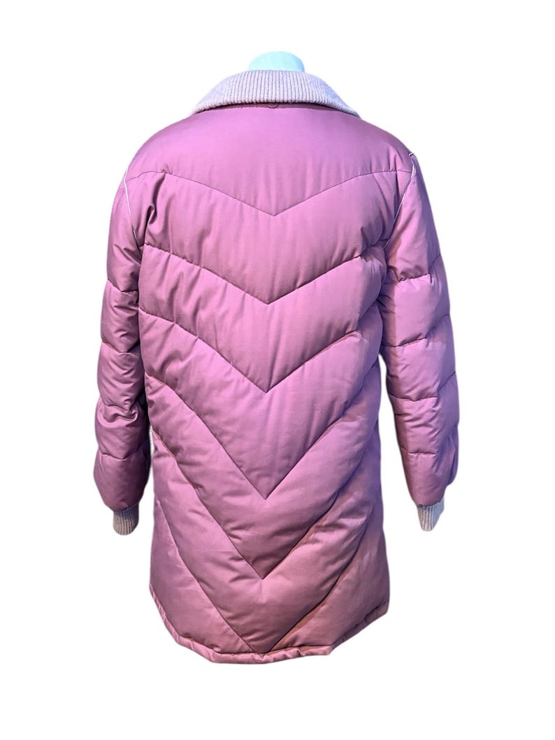 May include: A long, quilted, mauve-colored coat with a chevron design. The coat has a light gray knit collar and cuffs. The coat is displayed on a mannequin.