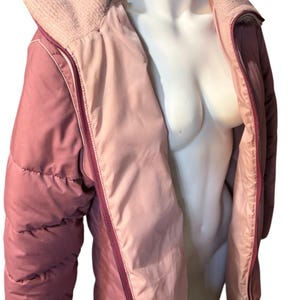 May include: A two-tone, quilted jacket in shades of pink and mauve. The jacket features a ribbed knit collar and a full-length zipper. The interior lining is a lighter shade of pink, creating a contrasting effect. The jacket is displayed on a mannequin.