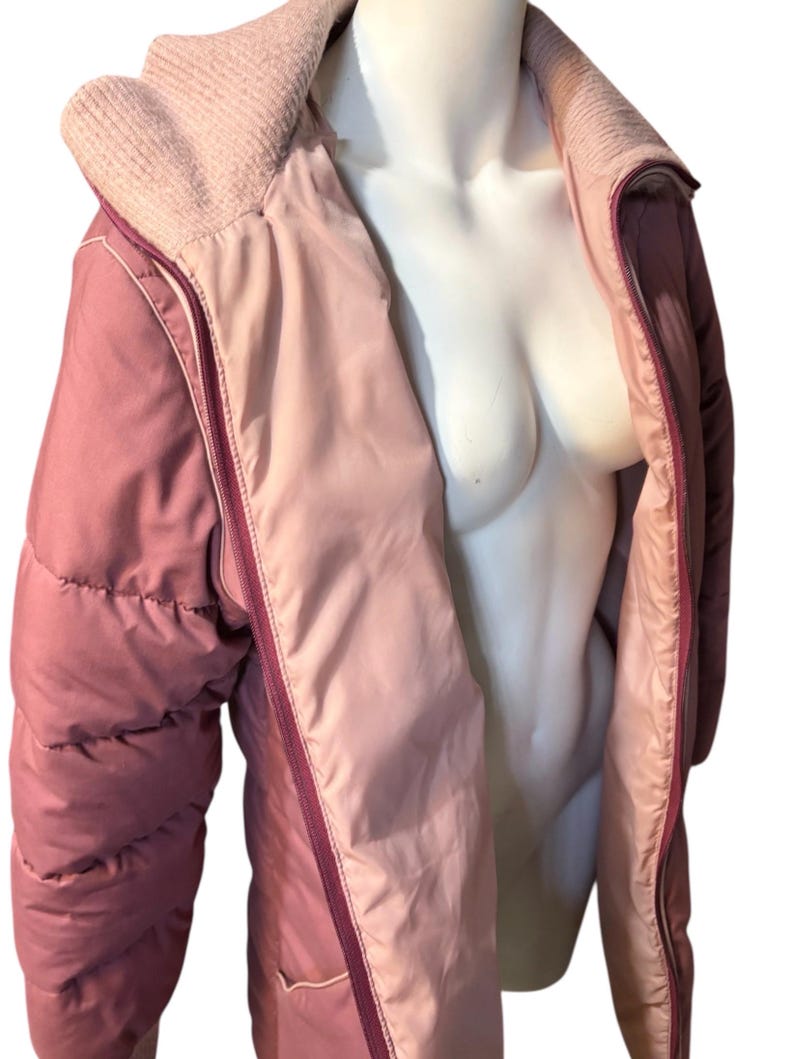 May include: A two-tone, quilted jacket in shades of pink and mauve. The jacket features a ribbed knit collar and a full-length zipper. The interior lining is a lighter shade of pink, creating a contrasting effect. The jacket is displayed on a mannequin.