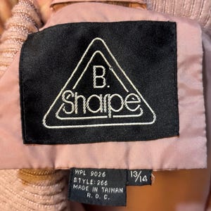 May include: Close-up of a pink jacket label with the brand name "B. Sharpe" in a white, triangular design on a black background. The label also includes size 13/14 and other text.