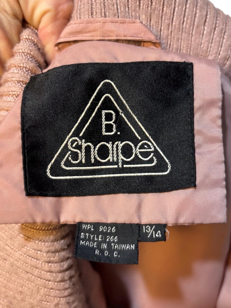 May include: Close-up of a pink jacket label with the brand name "B. Sharpe" in a white, triangular design on a black background. The label also includes size 13/14 and other text.