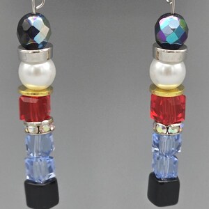 May include: A pair of dangling earrings with a silver hook. Each earring features a faceted, iridescent bead, a silver spacer, a white pearl bead, a gold spacer, red cube beads, clear crystal beads, blue cube beads, and a black square bead.