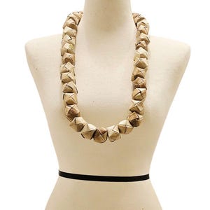 May include: A natural-toned necklace made of woven, geometric-shaped pieces. The necklace is displayed on a mannequin with a wooden neck and a black band around the waist. The necklace has a rustic, earthy aesthetic.