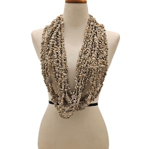 Puede incluir: Un collar de múltiples hilos hecho de pequeñas conchas marinas beige. El collar está drapeado alrededor de un maniquí, mostrando su diseño en capas. Las conchas tienen una apariencia natural y texturizada, creando una estética playera.