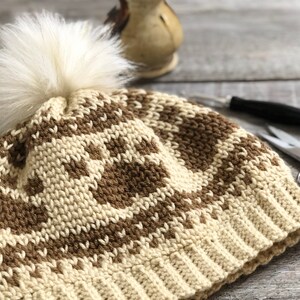 May include: A cream and brown knitted hat with a faux fur pom-pom. The hat has a repeating pattern and a ribbed brim. Scissors and a crochet hook are in the background.